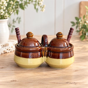 May include: A double ceramic condiment jar set featuring two connected pots with a two-tone brown and cream glaze. Each pot includes a matching removable lid and a small brown wooden spoon. A central handle connects the two rounded containers.
