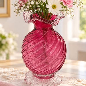 May include: A deep magenta glass vase with a swirling ribbed texture, a ruffled top rim, and a clear, textured, scalloped base. The vase is filled with a bouquet of white daisies, pink carnations, and small white baby's breath flowers.