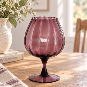 May include: A translucent purple glass vase with a wide, rounded bowl and a scalloped rim. The vase features vertical ribbed detailing along the body and sits on a slender stem with a circular base, crafted from smooth, polished purple glass.