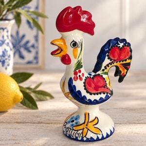 Pode incluir: Uma estatueta de galo em cerâmica com uma base branca brilhante e detalhes pintados à mão em vermelho, azul, amarelo e verde. O galo tem uma crista vermelha proeminente, um bico amarelo e padrões florais no corpo. A palavra Portugal está na base.