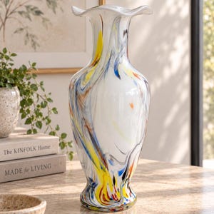 May include: A tall glass vase with a flared, ruffled rim. The body of the vase is primarily white with swirling, marbled streaks of yellow, blue, and orange throughout the glass. The base is slightly wider than the neck, creating a classic silhouette.