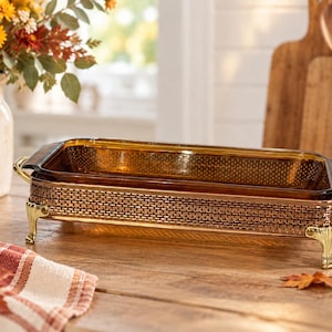 May include: A rectangular amber glass baking dish set inside a metallic gold-toned frame with a woven texture pattern. The frame features four ornate, curved legs and two side handles, providing a raised base for the glass insert.