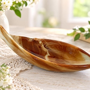 Peut inclure: Un bol décoratif en forme de feuille présentant une finition lisse et polie avec des motifs tourbillonnants dans des tons brun profond, ambre et blanc crème. Le récipient a une silhouette allongée et incurvée qui se termine en pointe.