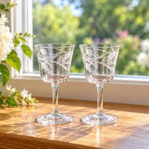 Könnte beinhalten: Zwei klare Weingläser mit Stiel, die mit zarten, eingravierten Distelmustern an den oberen Kelchen verziert sind. Jedes Glas hat eine glatte, transparente Oberfläche, einen schlanken Stiel und einen runden Fuß, was ein elegantes Design unterstreicht.