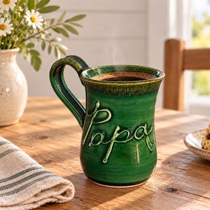 Peut inclure: Une tasse en céramique vert émeraude brillant avec un bord brun foncé et une grande anse. Le mot "Papa" est embossé en lettres cursives en relief sur le devant de la tasse. La tasse est remplie d'un liquide sombre et dégage une fine vapeur.