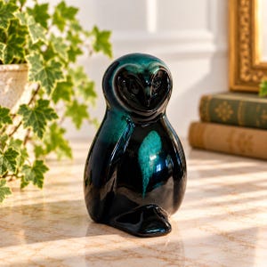 Puede incluir: Una figura de búho de cerámica con acabado brillante, color base negro profundo y acentos azul cerceta vibrantes en la cabeza y el pecho. El búho tiene un cuerpo liso y redondeado, un disco facial en forma de corazón y un pico puntiagudo.