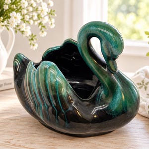 Puede incluir: Un macetero o cuenco de cerámica con forma de cisne que presenta un acabado brillante con una base de color negro intenso que se transforma en vibrantes reflejos verde esmeralda en las alas y el cuello, con una silueta curva y centro hueco.