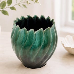 May include: A ceramic planter with a deep emerald green glaze and a dark, near-black base. The vessel features a rounded, bulbous body with vertical, swirling ridges that extend upwards to form a scalloped, petal-like rim with a glossy finish.