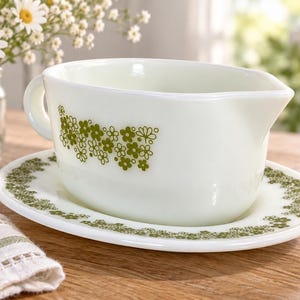 May include: A white milk glass gravy boat and matching oval underplate featuring a repeating green floral pattern. The gravy boat has a single handle and a pouring spout, while the oval plate has a matching green floral border design.