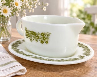 May include: A white milk glass gravy boat and matching oval underplate featuring a repeating green floral pattern. The gravy boat has a single handle and a pouring spout, while the oval plate has a matching green floral border design.