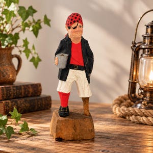 Puede incluir: Una figura de pirata tallada en madera sobre una base de bloque. El personaje lleva un pañuelo rojo con puntos negros, una chaqueta negra, una camisa roja y pantalones blancos. Tiene un parche en el ojo, una pata de palo y una taza gris.