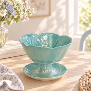 May include: A glossy, turquoise ceramic bowl with a scalloped, flower-like rim, supported by a pedestal base that rests on a matching circular plate. The surface features a reflective, iridescent glaze with subtle variations in colour depth.
