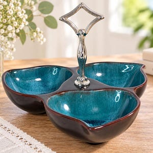 May include: A three-section serving dish featuring a glossy, deep teal interior glaze that transitions into a dark brown exterior. The ceramic bowls are connected at the centre by a polished silver-tone metal handle with a diamond-shaped top loop.