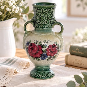 May include: A ceramic vase featuring a cream-coloured body with hand-painted red roses and blue accents. The vase has a dark green glazed neck and base with intricate embossed scrollwork patterns and two side handles for a classic aesthetic.