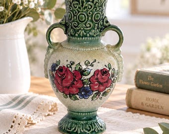 May include: A ceramic vase featuring a cream-colored body with hand-painted red roses and blue accents. The vase has a dark green glazed neck and base with intricate embossed scrollwork patterns and two side handles for a classic aesthetic.