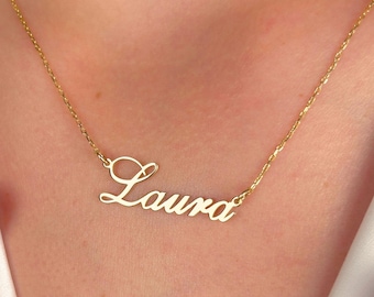 May include: A gold-colored necklace featuring a delicate chain and a central pendant in a cursive script font that spells out the name Laura. The metal has a polished finish, and the pendant is attached to the chain on both sides of the name.