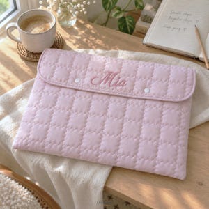 May include: A light pink quilted laptop sleeve featuring a rectangular flap with two white snap buttons and the name Mia embroidered in a cursive script on the front. The sleeve has a soft, grid-like stitched texture across the entire surface.