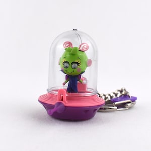May include: A keychain featuring a small green alien figure with pink antennae inside a clear plastic dome. The base is a pink and purple saucer-shaped vessel with a small purple antenna protruding from the front, attached to a silver metal chain.