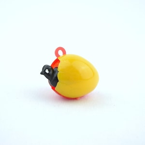 Puede incluir: Un pequeño amuleto en forma de huevo amarillo con un borde rojo y un pequeño lazo rojo en la parte superior. Un pájaro negro emerge del huevo. El amuleto está sobre un fondo blanco.