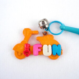 Op de afbeelding: Oranje scooter-vormige sleutelhanger met het woord "FUN" in kleurrijke letters. De sleutelhanger heeft een zilveren bel en een blauwe sluiting. De scooter heeft rode en oranje accenten.