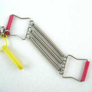 Può includere: Un attrezzo per esercizi a molle metalliche con tre molle di tensione argentate parallele collegate a due telai in filo metallico con impugnature in plastica rossa. Una piccola campanella argentata e un anello di plastica gialla sono fissati.