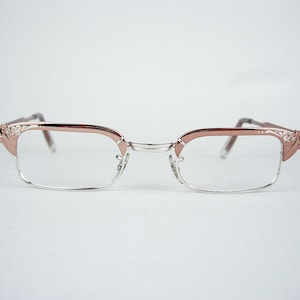 May include: Rose gold and silver framed eyeglasses. The glasses have a half-rim design with clear lenses. The temples are a darker colour, and the corners of the frames have decorative detailing.