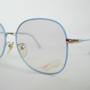 Puede incluir: Gafas de vista azul claro con montura de metal dorado y lentes transparentes. El nombre de la marca "Tura" está impreso en la lente derecha.