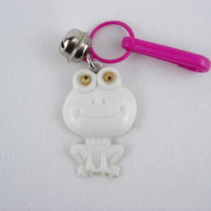 May include: A white frog-shaped keychain with gold-colored eyes and a smiling face. It has a silver bell and a pink plastic clip. The keychain is designed to attach to keys or bags.