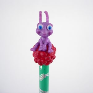 Könnte beinhalten: Eine lila Ameisenfigur mit blauen Augen sitzt auf einer roten, beerenartigen Struktur. Die Figur ist an einem grünen Bleistift mit weißen und silbernen dekorativen Elementen befestigt.