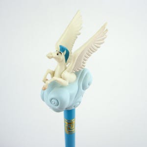 以下が含まれることがあります： ペガサスのフィギュアが付いた青い鉛筆。ペガサスは白で、クリーム色の翼と青いタテガミがあります。鉛筆の消しゴムは水色の雲です。