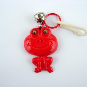 May include: A red frog-shaped keychain with a silver bell and a white clip. The frog has large, golden eyes and a smiling mouth. The keychain is attached to a red ring.