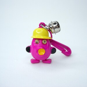 May include: A pink plastic toy with a yellow hard hat, googly eyes, and an open mouth. The toy has a silver bell attached to it and a plastic clip.