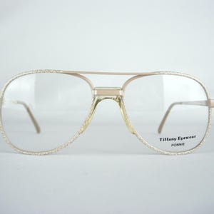 May include: Gold-toned aviator eyeglasses with clear lenses. The frames have a textured, glittery border and a double bridge. The text "Tiffany Eyewear RONNIE" is printed on the lower right lens.