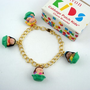 May include: Gold-tone charm bracelet featuring Cabbage Patch Kids charms. The bracelet has a chain link design with several colorful doll charms. A small box with the words "Cabbage Patch Kids Charm Bracelet" is also in the image.