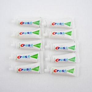 以下が含まれることがあります： 白い表面に配置された、Crest歯磨き粉の小さなチューブが10本。各チューブは白で、ブランド名「Crest」が赤と青で、緑の三角形が付いています。チューブは旅行用または試用です。