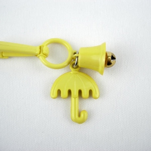 May include: A yellow plastic charm with a bell and umbrella design. The charm has a clip, a ring, a bell, and an umbrella pendant. The bell is gold-colored, and the umbrella is yellow. The charm is on a white background.