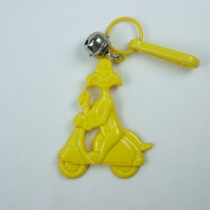 Puede incluir: Llavero de plástico amarillo con un gato de dibujos animados conduciendo un scooter. El llavero tiene una campana plateada.