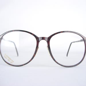 May include: Round-framed eyeglasses with a tortoiseshell pattern. The frames are a mix of brown and black, with clear lenses. The text "CARBON NO-SLIP" is printed on the lower left lens. The temples are thin and silver.