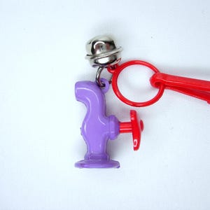 May include: A purple tap-shaped keyring with a red handle and a silver bell attached. The keyring has a red plastic clip and ring. The background is plain white.