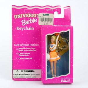 Può includere: Un portachiavi Barbie University nella sua confezione rosa originale. Il portachiavi presenta una bambola cheerleader con i capelli biondi, che indossa un'uniforme arancione e bianca. La confezione include testo sulle caratteristiche della bambola e un avvertimento di pericolo di soffocamento.
