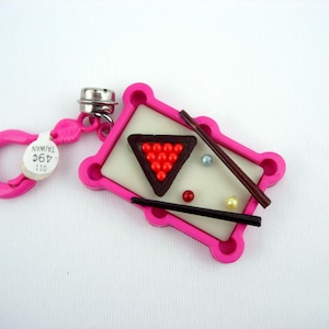 May include: A miniature pink pool table keychain with a white playing surface, a red triangle of billiard balls, and two cues. The keychain includes a pink clasp and a tag that reads "49¢ TAIWAN".