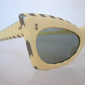 Puede incluir: Un par de gafas de sol vintage de ojo de gato con montura de color crema y rayas negras. Las gafas tienen lentes grises y un pequeño diseño de estrella en la montura.