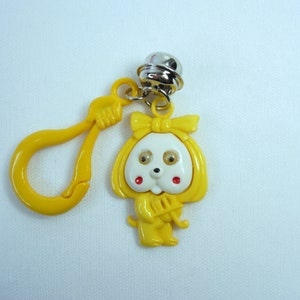 May include: A yellow plastic dog charm with a bow, red cheeks, and a silver bell. The charm has a yellow clasp and is on a white background. The dog is holding a musical instrument.