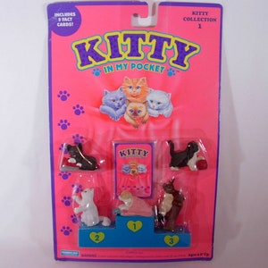 Könnte beinhalten: Eine pinkfarbene und violette Kartenverpackung für eine Kitty in My Pocket Spielzeugsammlung. Die Verpackung zeigt ein Bild von vier Kätzchen in einem rosa Bett. Die Verpackung enthält fünf Faktenkarten und drei kleine Plastik-Kätzchenfiguren. Der Text auf der Verpackung lautet "Kitty in My Pocket" und "Kitty Collection 1".