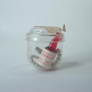 Puede incluir: Un pequeño recipiente de plástico transparente con una botella roja en su interior. La botella tiene una etiqueta blanca que dice "Grease Stud".