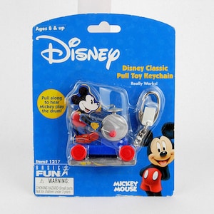 Può includere: Un portachiavi Disney Classic Pull Toy con Topolino che suona la batteria. Il portachiavi è su un supporto di cartone blu con il logo Disney e il testo "Ages 8 & up". La confezione include anche il testo "Pull along to hear Mickey play the drum!"