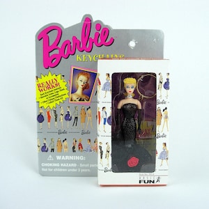 Può includere: Una bambola portachiavi Barbie in un abito nero con una rosa rossa, esposta in una scatola. La scatola è accanto a un supporto di cartone con il marchio Barbie. La confezione include il testo "REALLY WORKS!" e "BASIC FUN".