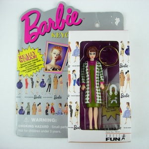 Può includere: Una bambola portachiavi Barbie in una scatola. La bambola indossa un cappotto verde e bianco fantasia sopra un vestito verde. La confezione include il logo Barbie, le parole "REALLY WORKS!" e "BASIC FUN".