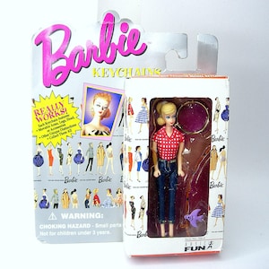 Può includere: Un portachiavi Barbie con una camicia a quadri rossi e bianchi, jeans blu e un ciondolo a forma di pesce viola. Il portachiavi è confezionato in una scatola bianca con le parole "Barbie Keychains" e "Really Works!" stampate su di essa.