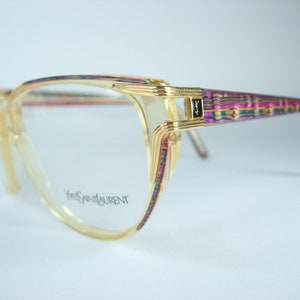 Puede incluir: Gafas de oro con montura transparente y ribetes con estampado rosa, verde y dorado. El nombre de la marca "Yves Saint Laurent" está impreso en la montura.